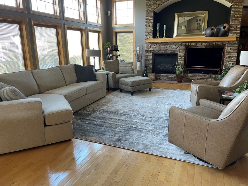 After design image of living room space with large windows and fire place. Milford two piece sectional with cuddle chaise and Louise Cuddle Chair and ottoman on left side of image. Stephanie swivel gliders in leather on right side of image. Rug pulls the whole room together.